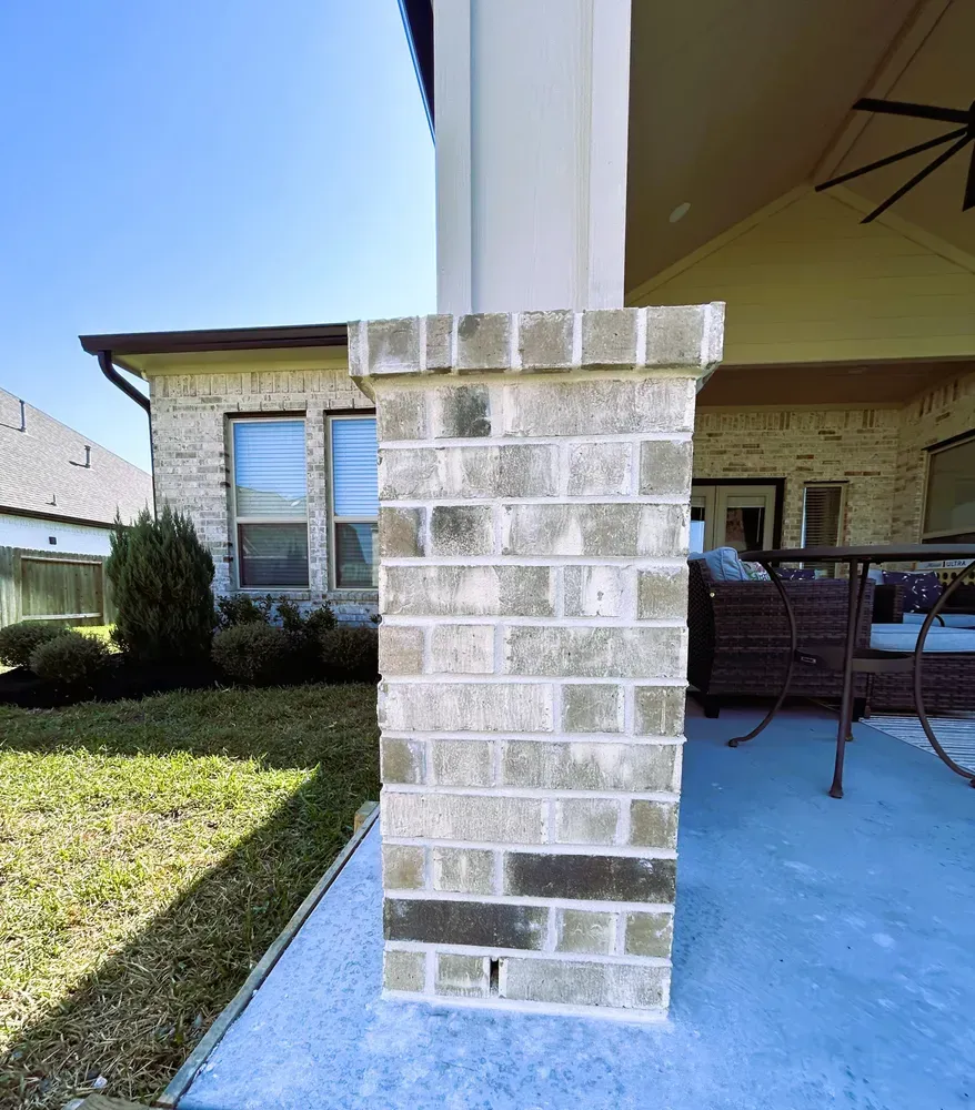 Brick column supporting a porch roof, light-colored bricks with a darker row. Grass and house in the background.
