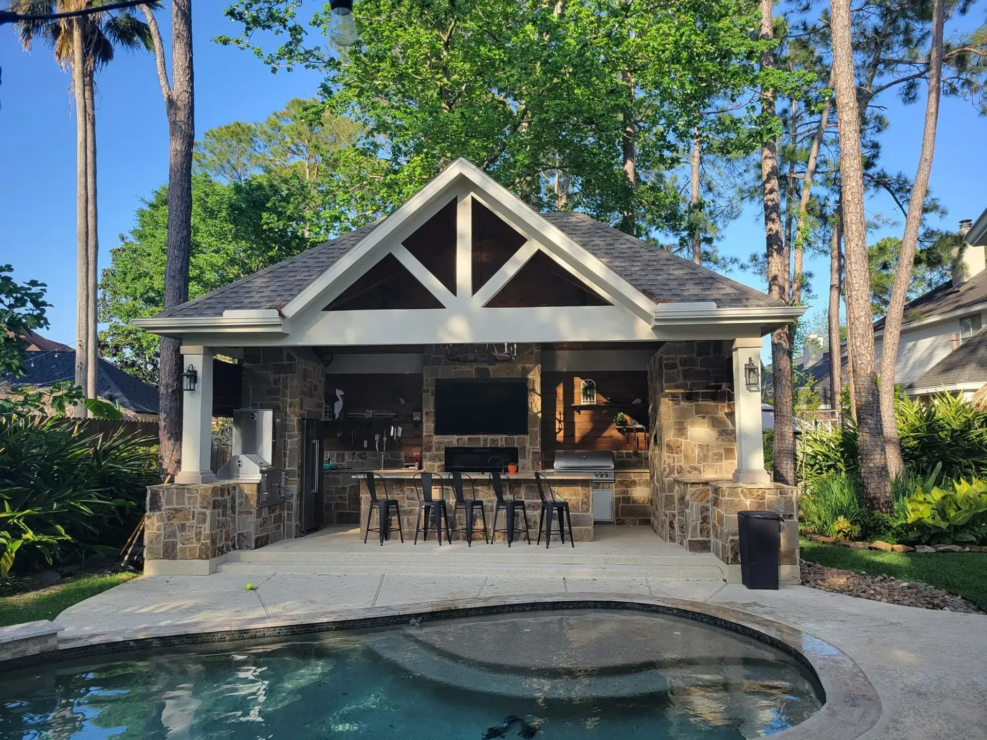 Outdoor kitchen with a pool, brown stone, bar, and TV.