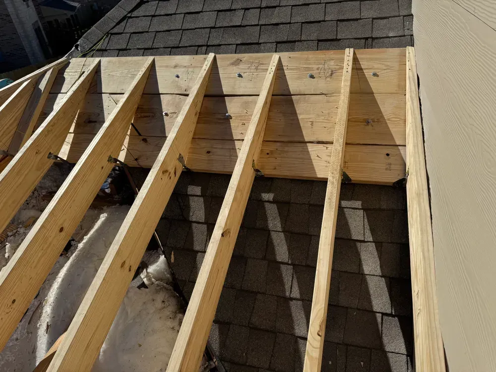 Wooden roof frame construction with exposed rafters, partially over dark shingles.
