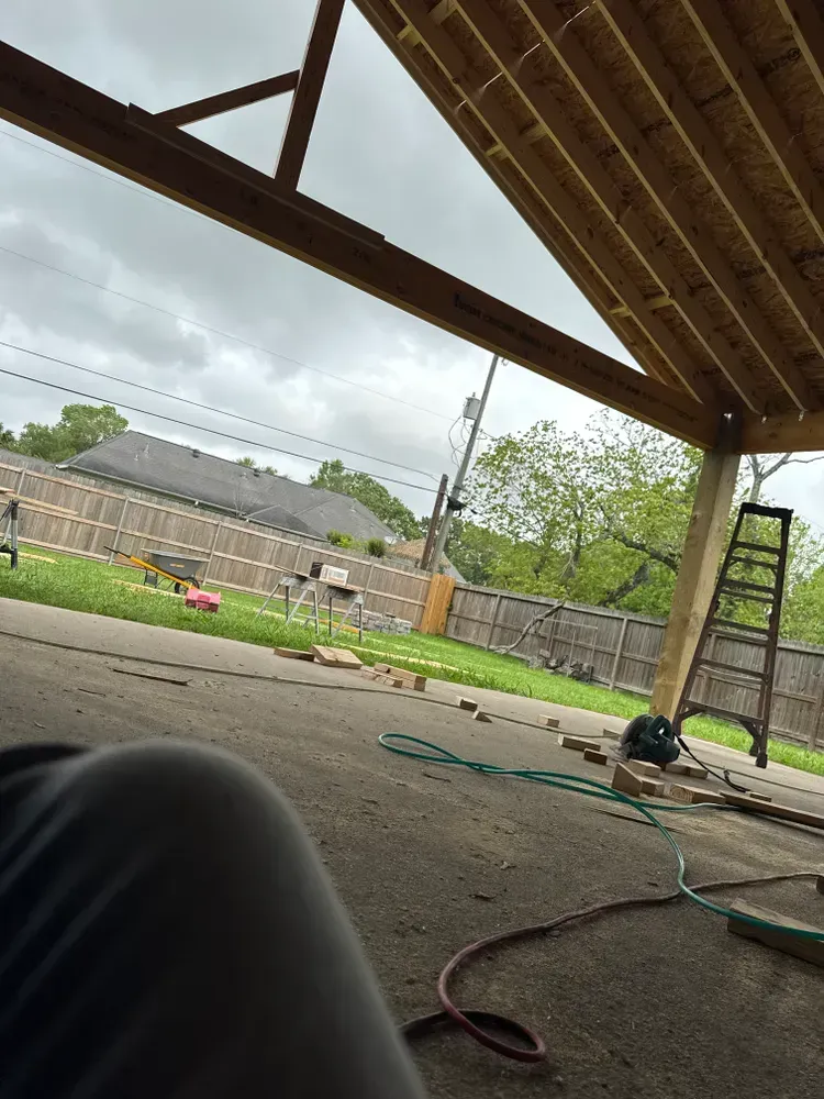 Patio roof under construction, with a ladder, tools, and backyard view. Cloudy sky.