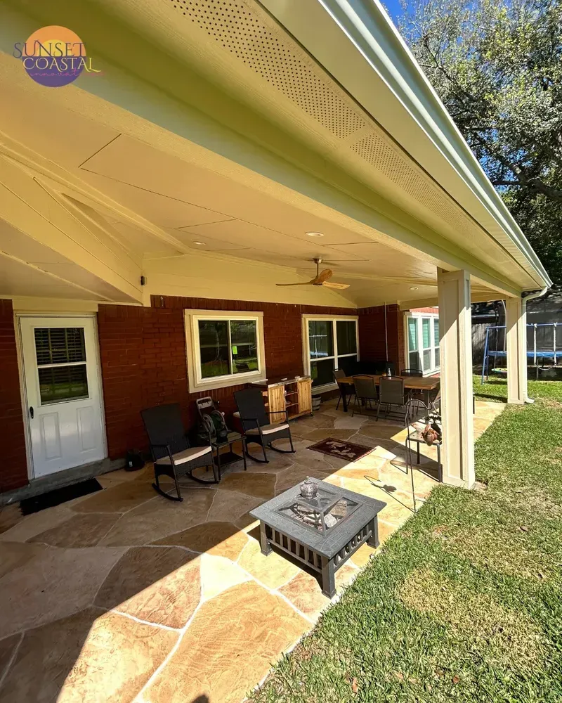 Covered patio with rock flooring, chairs, and fire pit next to grass and a red brick house.