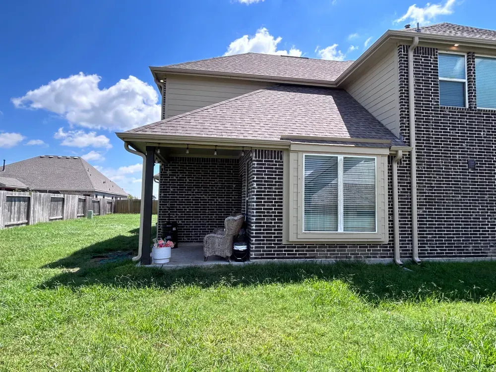 Backyard of a brick house with a covered patio; green grass, blue sky, and wooden fence in the distance.