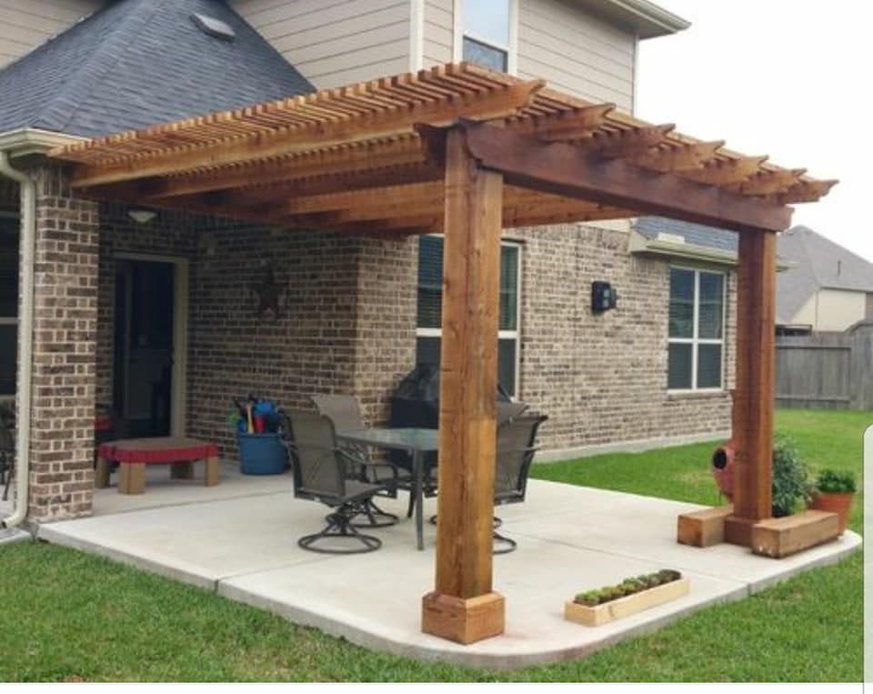 Wooden pergola over a concrete patio with a table and chairs, next to a brick house.