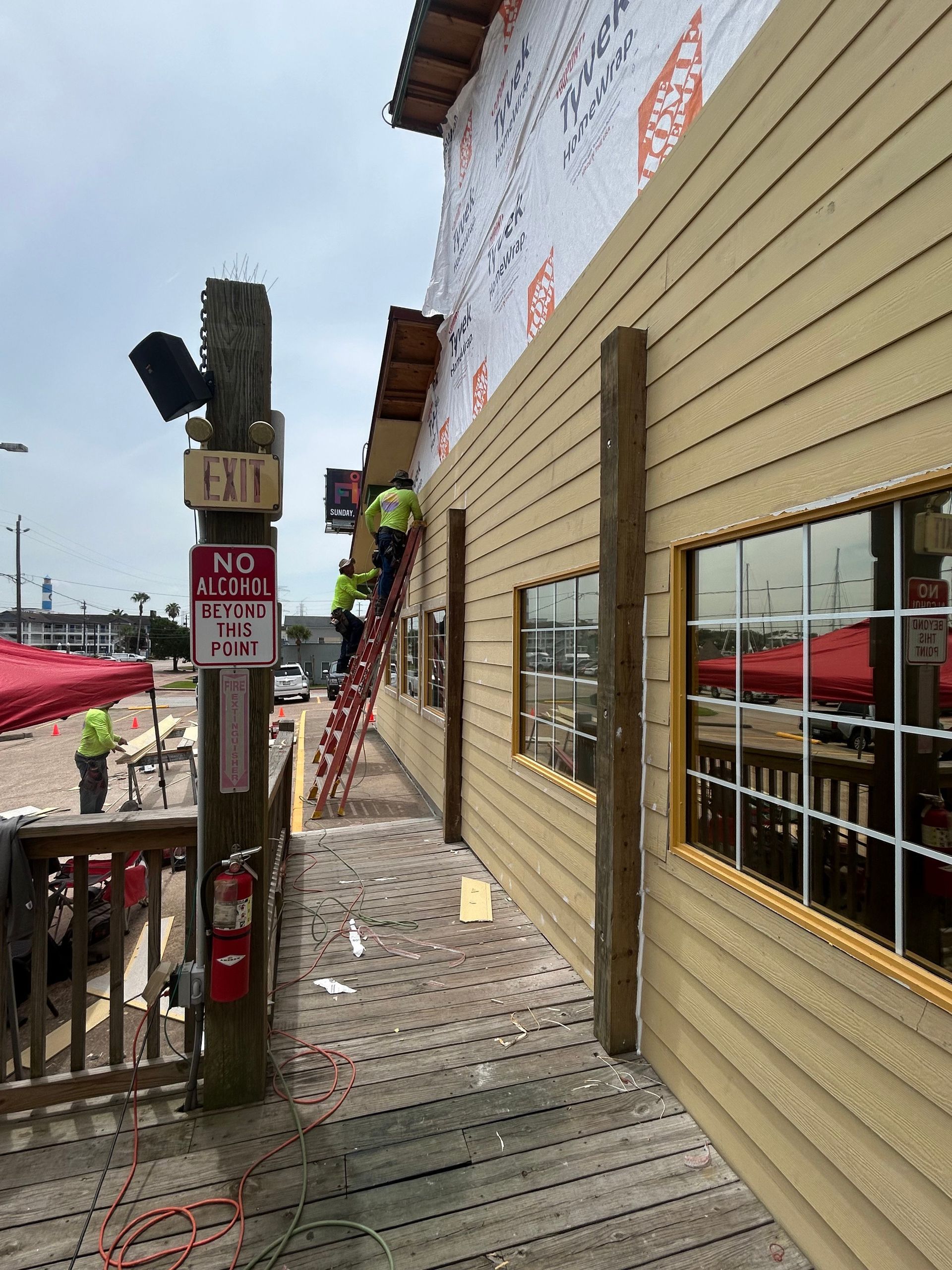 Construction workers installing siding on a restaurant. Yellow siding, brown posts, and a wooden deck are visible.