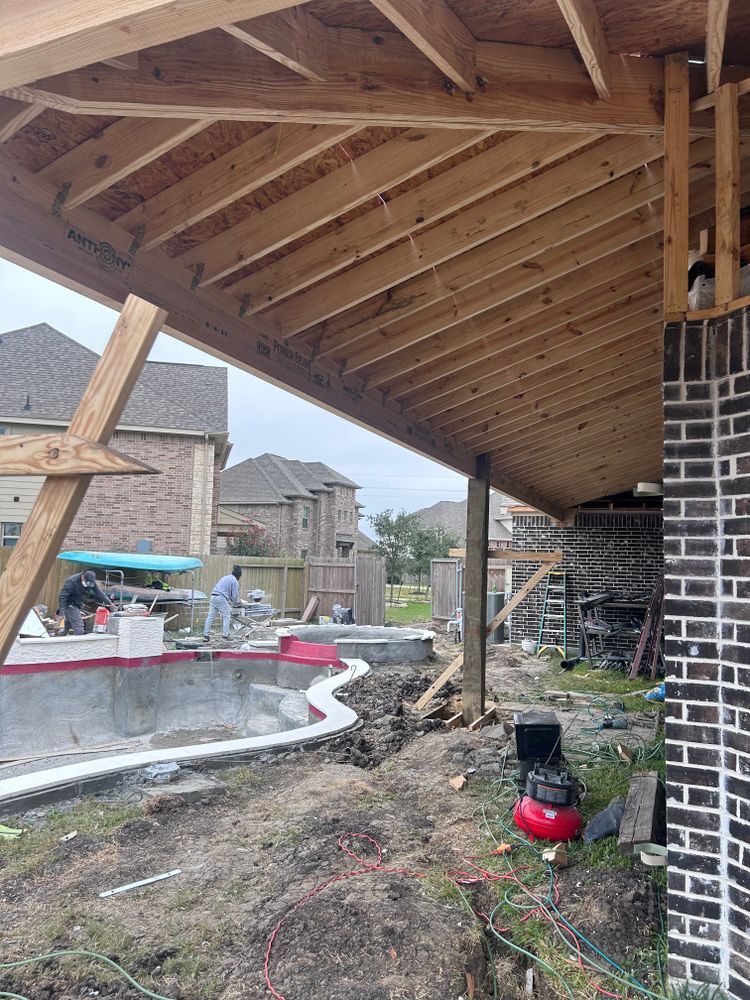 Construction of a covered patio, pool, and backyard. Workers are present. Brick chimney on right.
