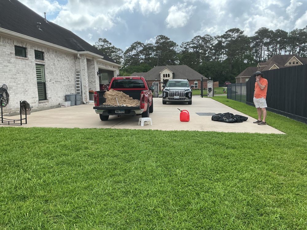 A red truck loaded with wood, a person, and a car on a concrete driveway next to a house and green lawn.
