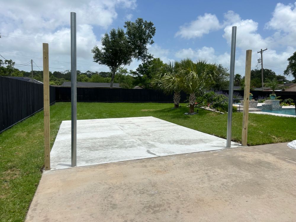 Concrete patio with four vertical poles set up in a backyard; greenery and a pool visible.