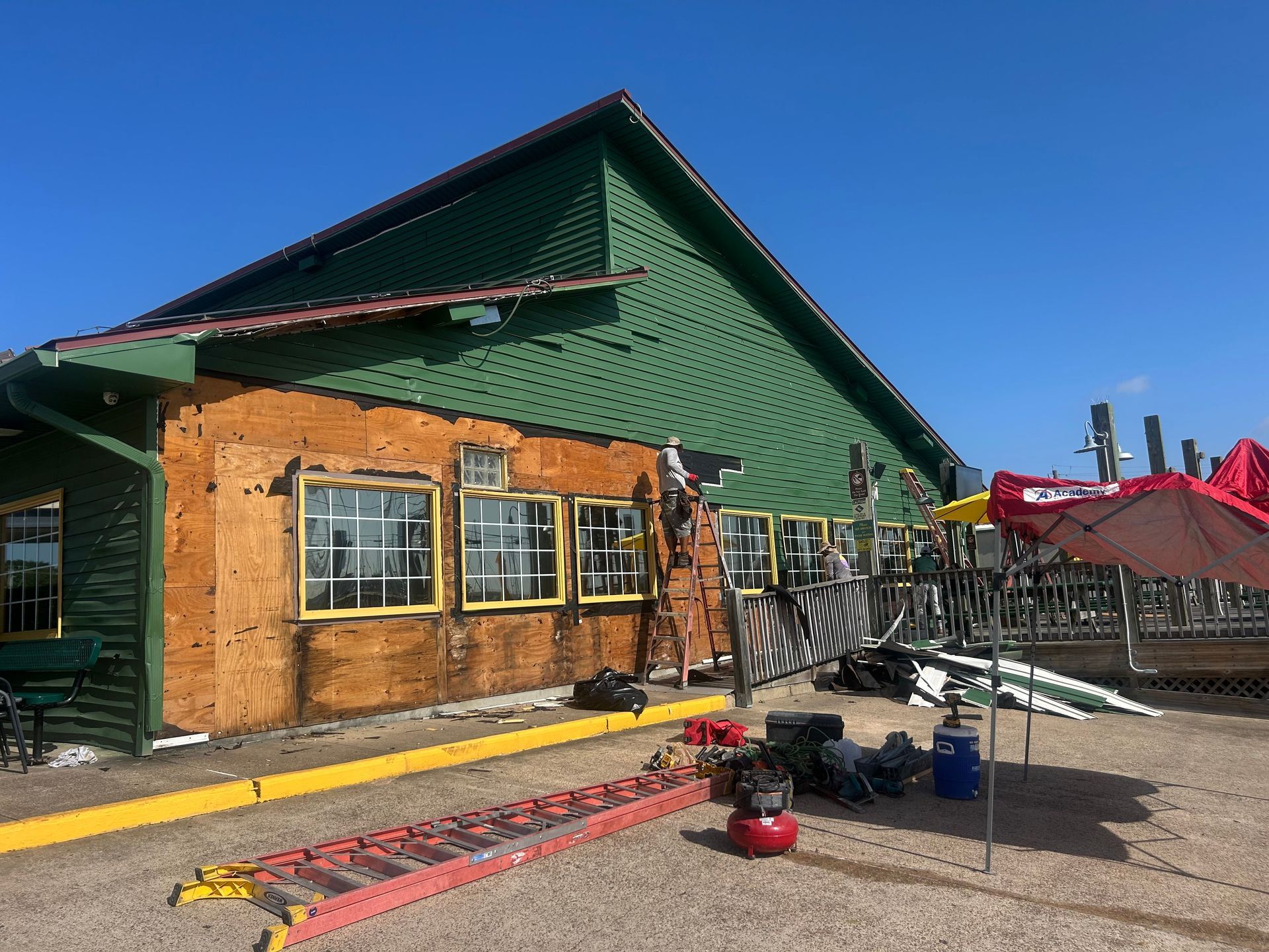 Building with green and brown siding being worked on; ladders, tools, and umbrella present.
