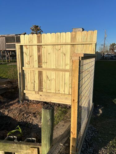 Wooden fence with picket top, built on a grassy patch near a body of water and houses.
