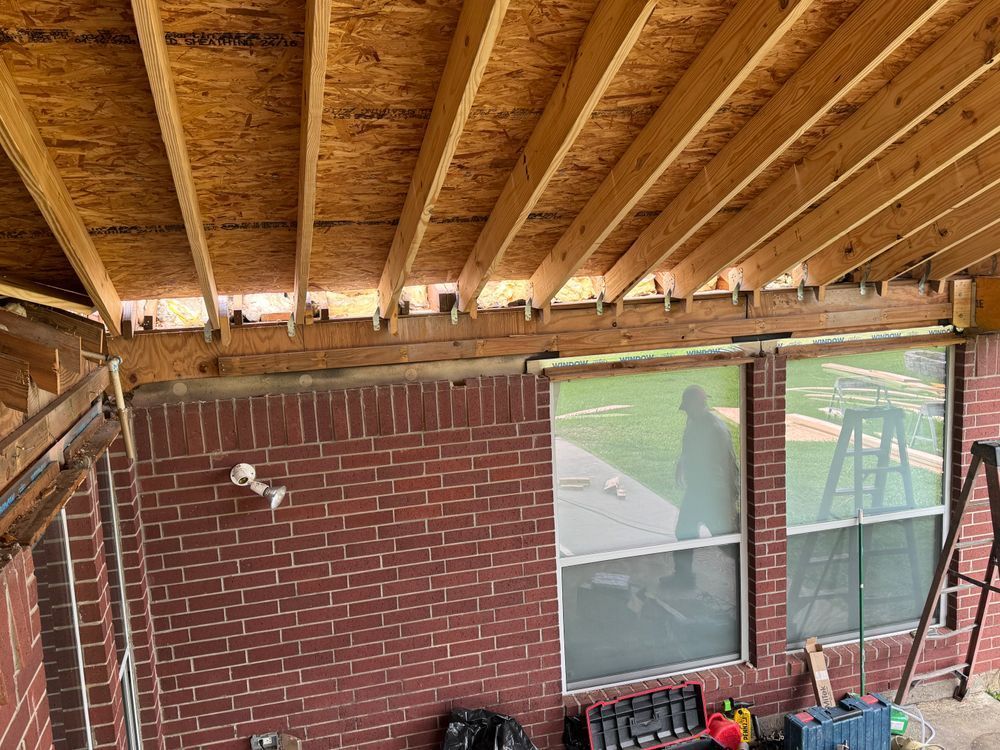 Construction of a wooden roof overhang over a brick wall, with visible framing and windows.
