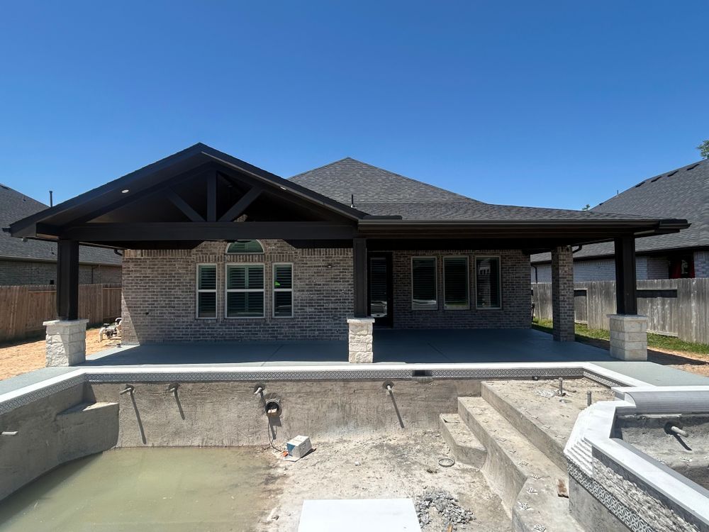 A house with a pool under construction. Brick facade, dark roof, and wooden covered patio. Sunny day.