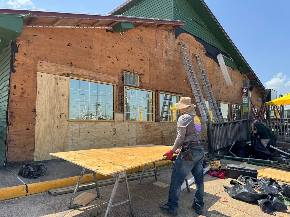 Construction worker installs plywood on the side of a building with windows. A-frame ladder and tools nearby.