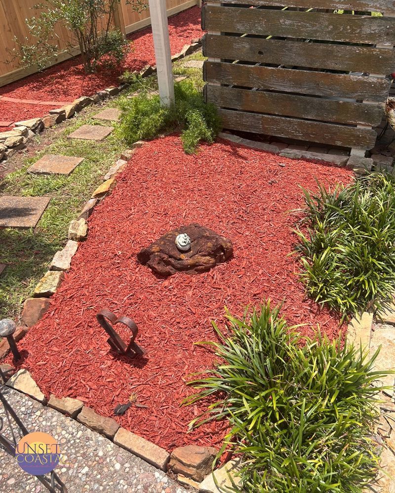 Red mulch garden bed with a decorative rock, bordered by brick and grass, with green plants and a wooden fence.