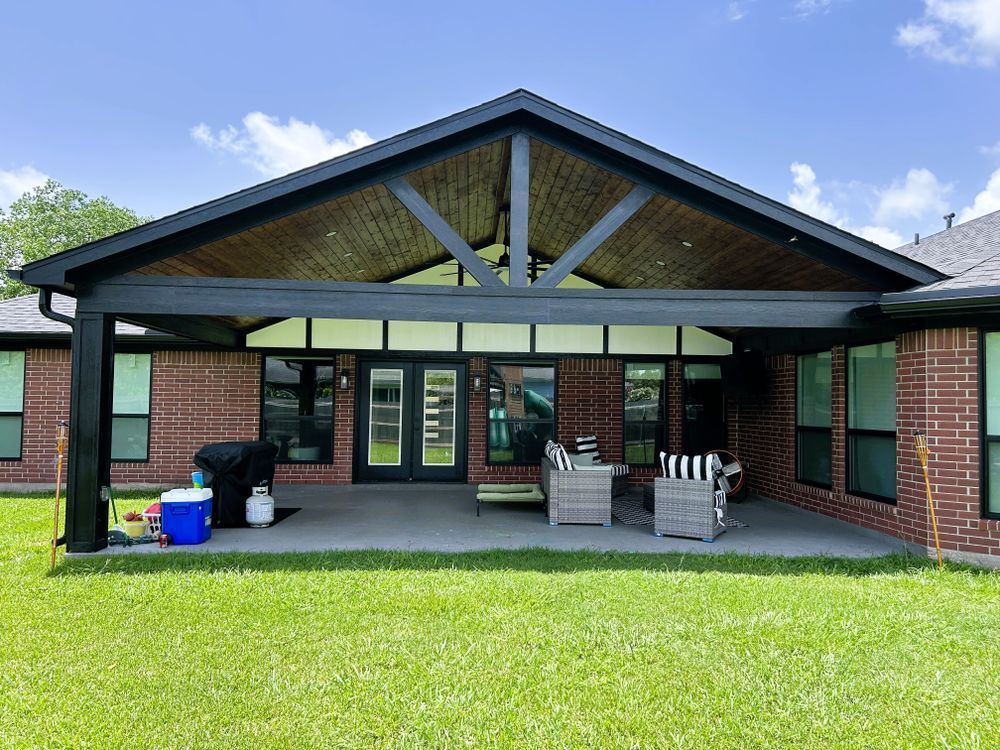 Covered patio with black frame, wood ceiling, brick house, outdoor seating on a concrete patio, and a grill.