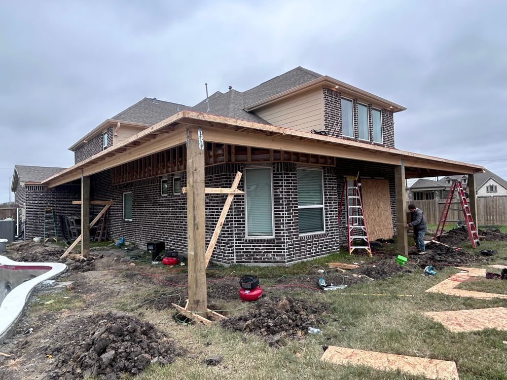 Construction of a covered patio on a two-story house; wooden frame and siding installation in progress.