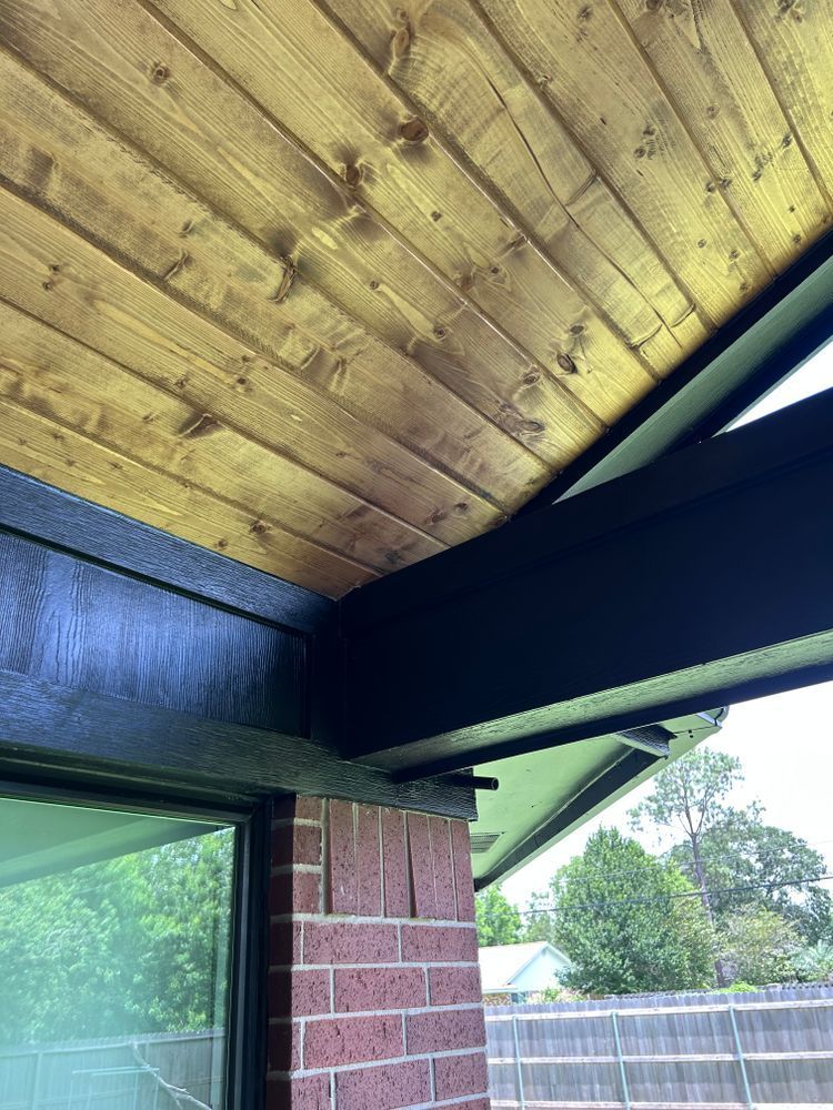 Wooden ceiling with dark beams, brick wall, and window frame, with a view of trees outside.