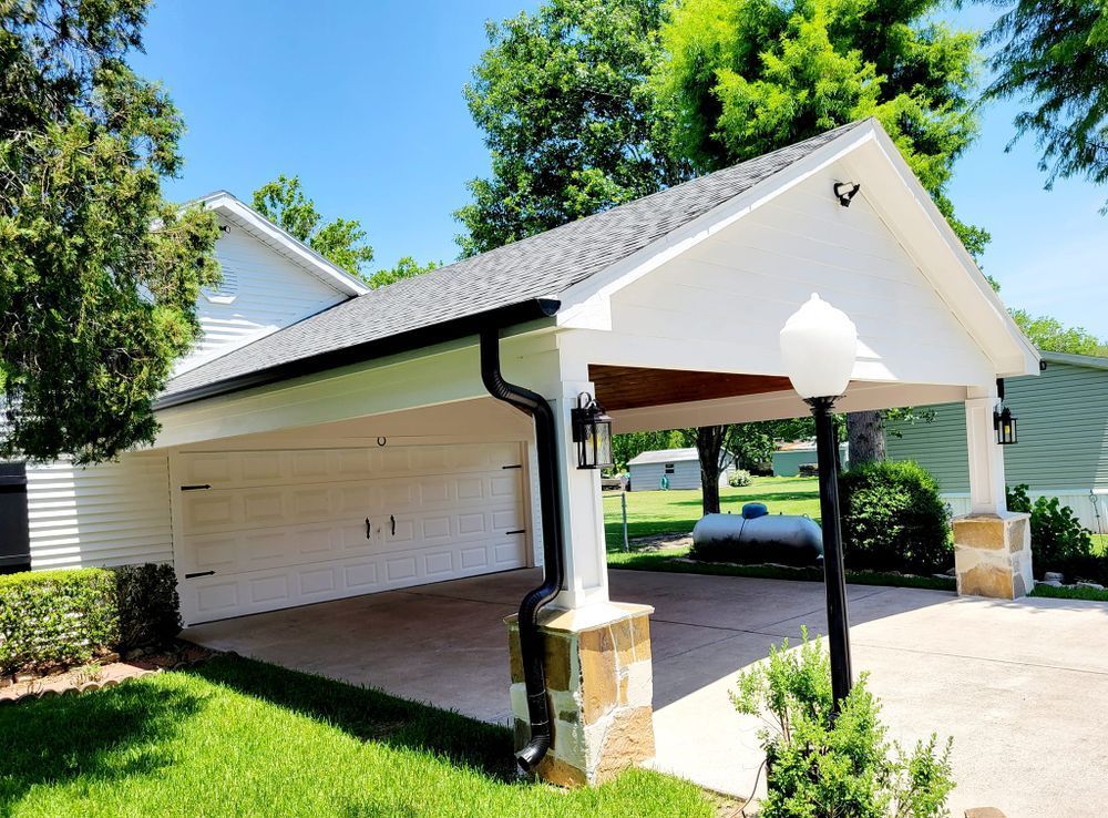 White carport with stone pillars and black roof, in a sunny green yard.
