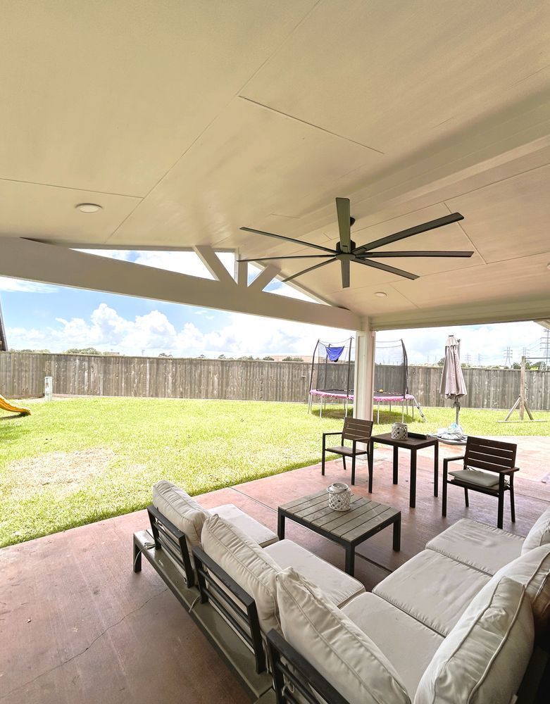 A covered patio with seating overlooking a grassy yard, fence, and blue sky.