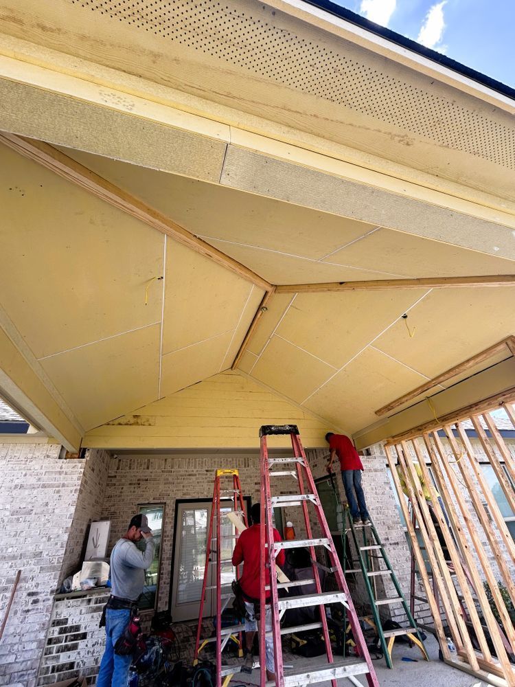 Workers installing drywall on a porch ceiling, with ladders, brick, and wooden framing.
