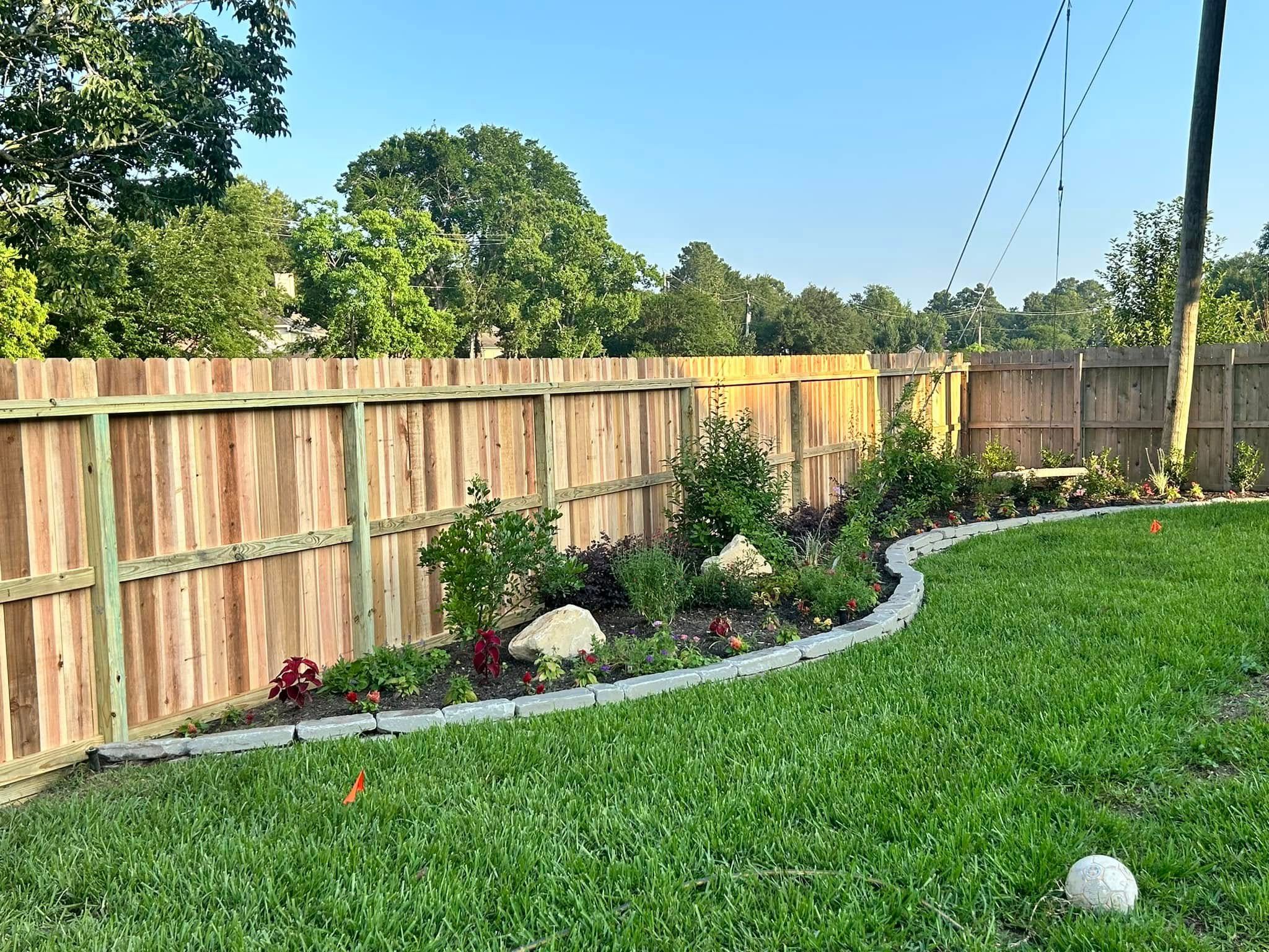 Wooden fence bordering a green lawn and a flower bed with rocks and various plants.