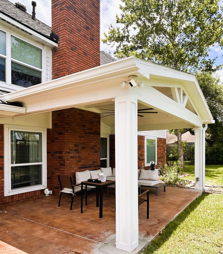 Covered patio with seating, attached to a brick house. White beams and columns, green lawn.