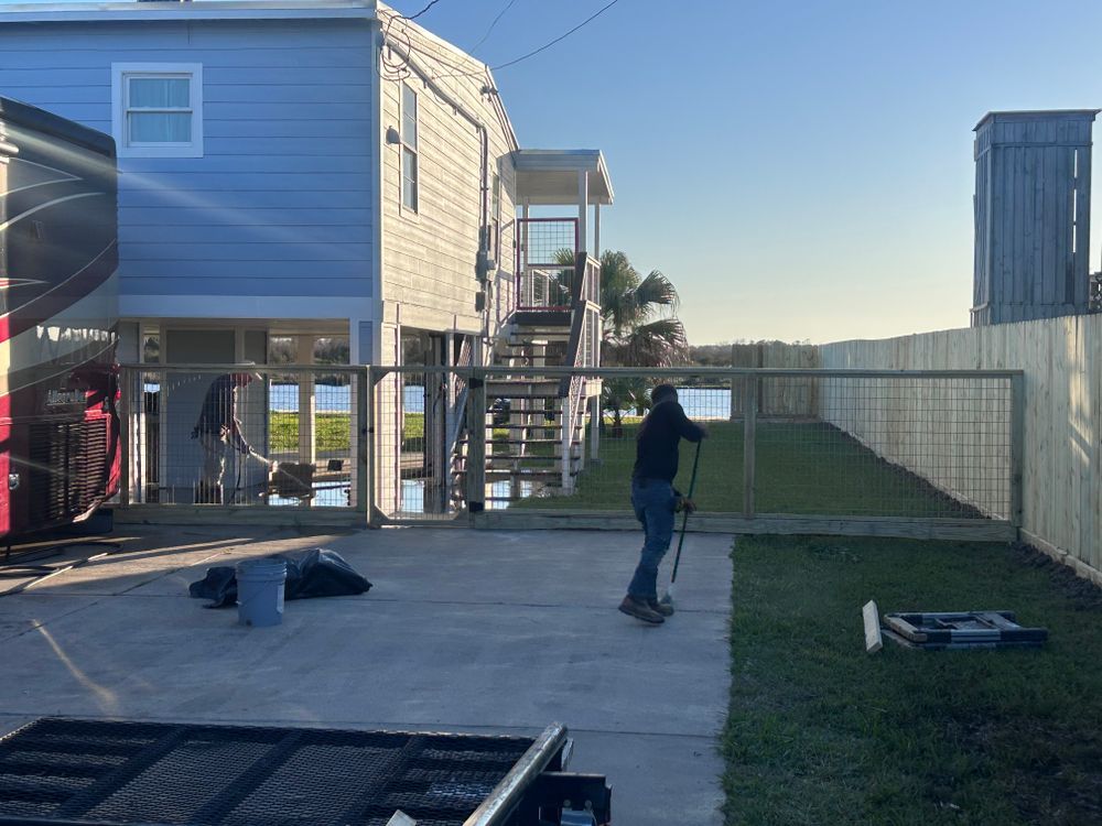 A person stands in a yard near a light blue house with fencing, sunshine, and a trailer.