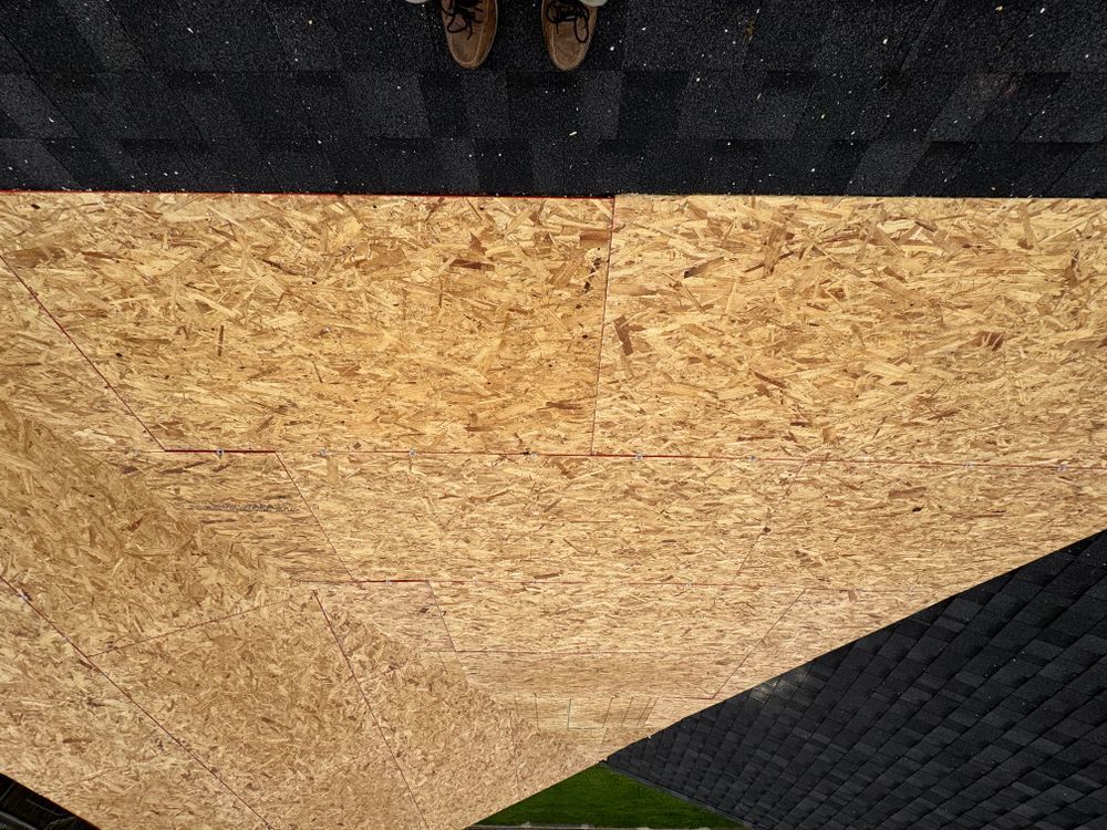 View from above of a roof under construction: plywood being installed, with worker's feet and existing shingle roof visible.