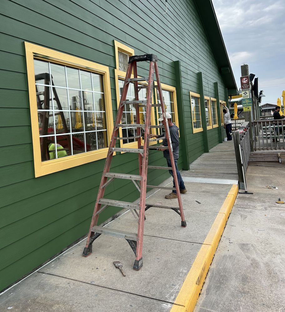Person on a ladder outside a green building with yellow-framed windows. Tools sit on the ground.