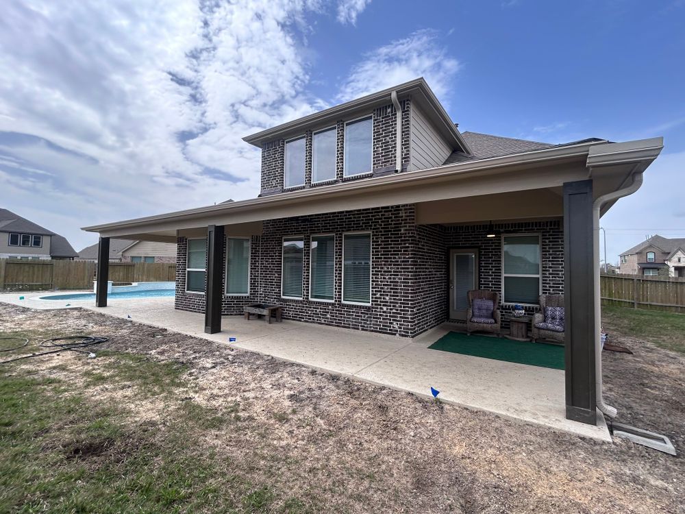 Backyard patio with a covered area, pool, and brick home under a blue sky.