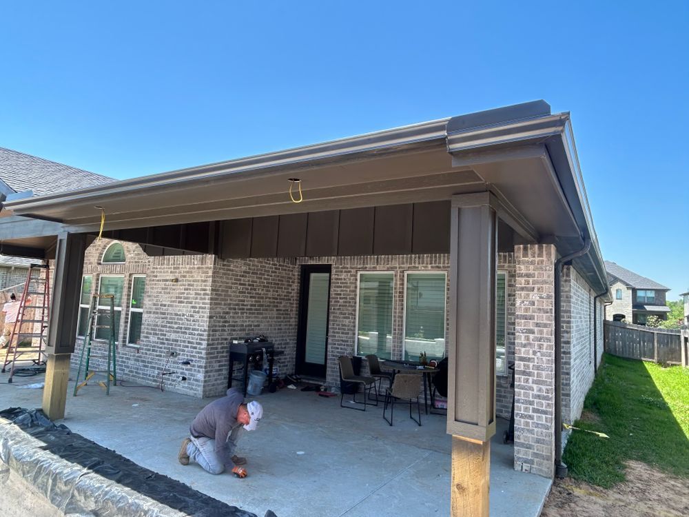 Patio under construction: Worker on hands and knees, brick house, brown awning, clear blue sky.