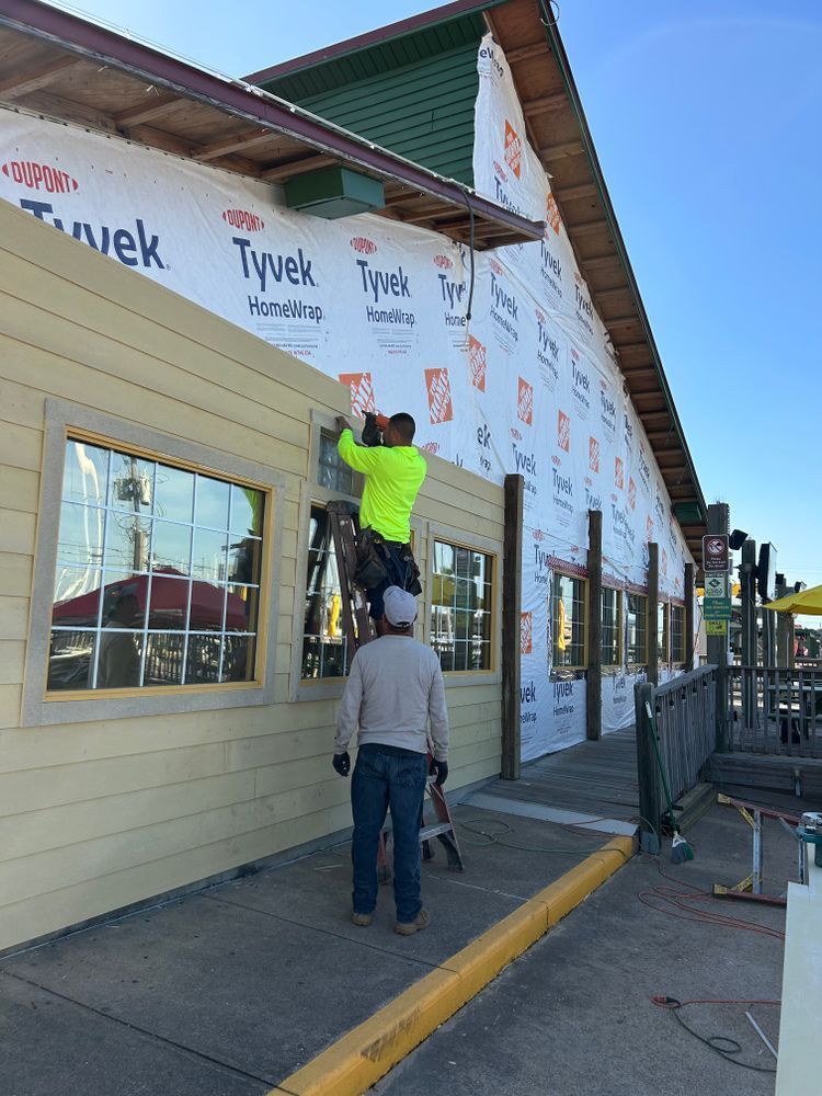 Two construction workers installing siding on a building with large windows, outdoors. One on a ladder.