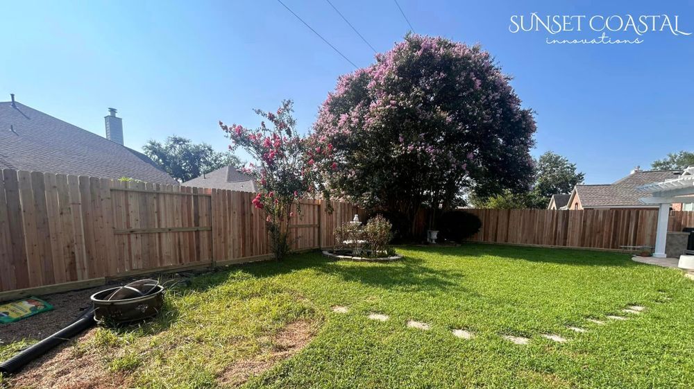 Backyard with wooden fence, green grass, and a large tree under a blue sky.