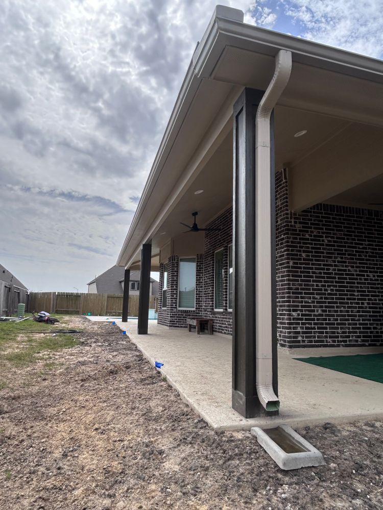 Covered patio with dark columns and a tan roof, concrete floor, and brick wall under cloudy sky.