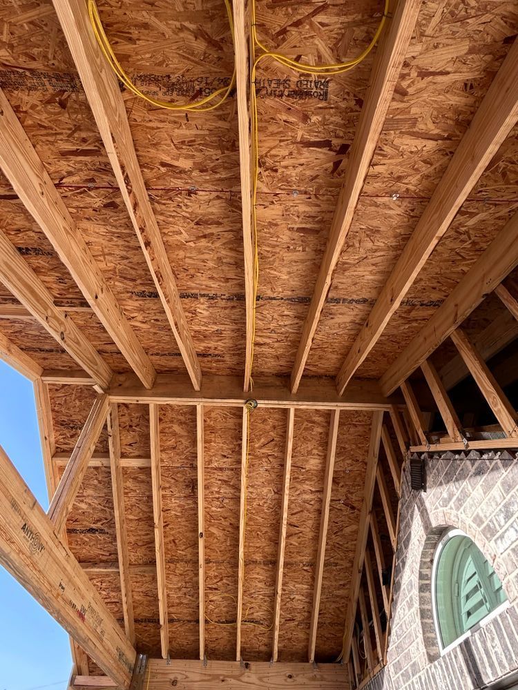 Wooden roof construction with OSB sheathing, electrical wiring, and partial view of a brick wall with an arched window.
