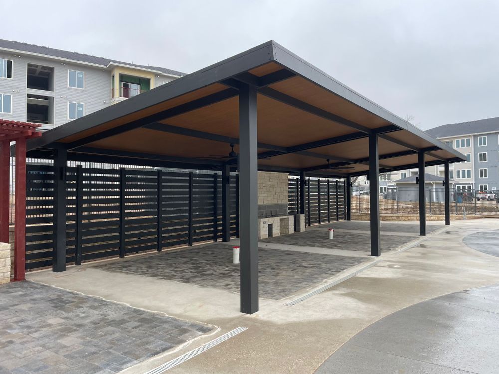 Black metal outdoor shelter with brown ceiling, brick floor, and wooden fence.