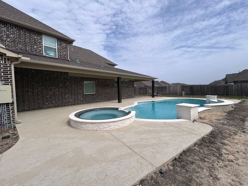 Backyard with a pool and spa, concrete patio, and brick house under a cloudy sky.