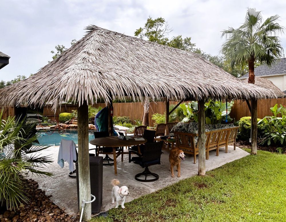 A thatched roof patio with a pool, seating, a person, and two dogs in a backyard setting.
