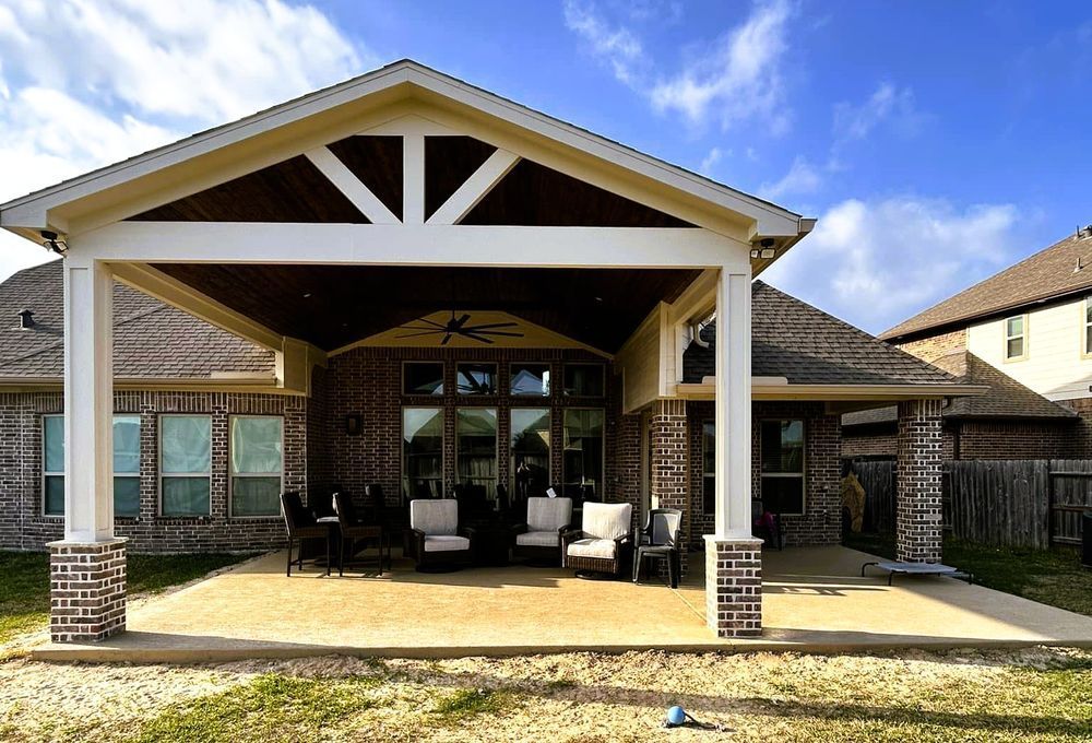 Covered patio with seating area, brick columns, and a brown ceiling against a house and cloudy sky.