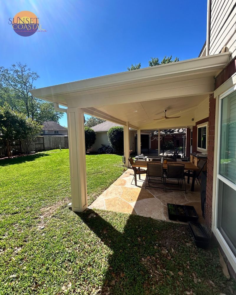 A covered patio with a dining set, adjacent to a house. Green grass and a sunny blue sky.