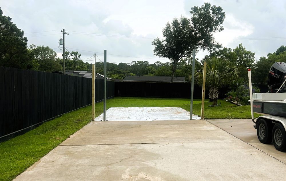Concrete driveway with boat trailer. Wooden poles mark a rectangular area. Green grass and trees in the background.