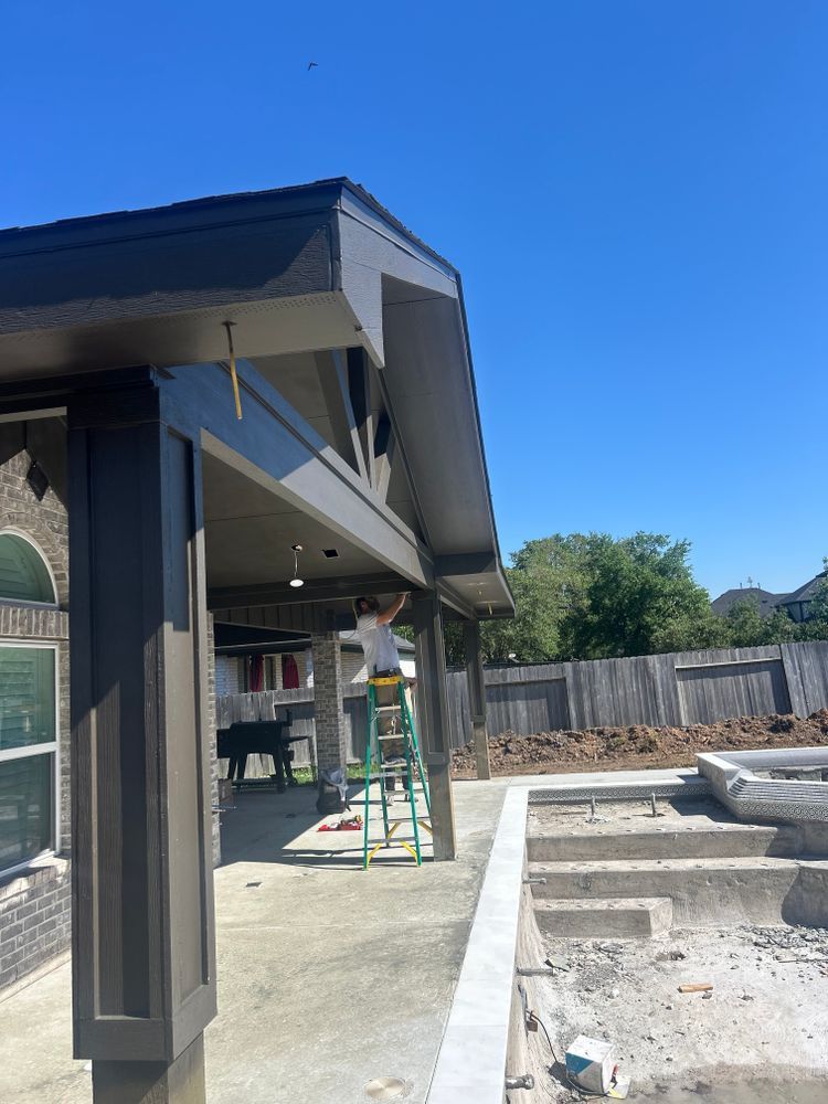 Construction worker on a ladder installing a light fixture on a patio with a dark roof; pool in the background.