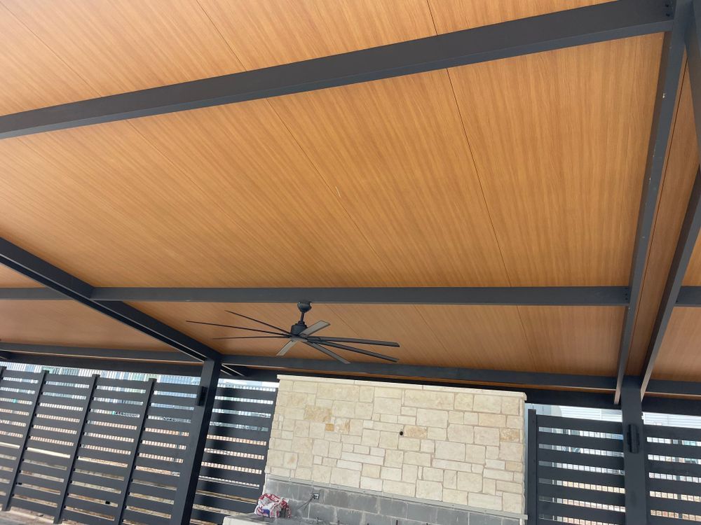 Brown ceiling with black beams and a ceiling fan over a patio with a stone wall and slatted fence.