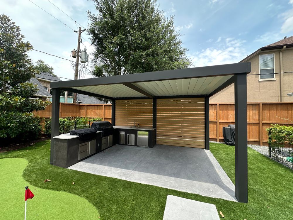 Outdoor kitchen under a pergola with grill, cabinets, wooden slatted wall, and putting green on grass.