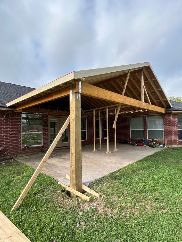 Construction of a wooden patio cover attached to a brick house, with a partially built roof and support beams on a concrete patio.