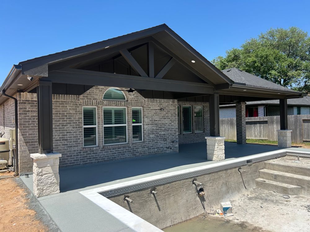 Brick house with dark brown pergola, empty pool, and blue sky.