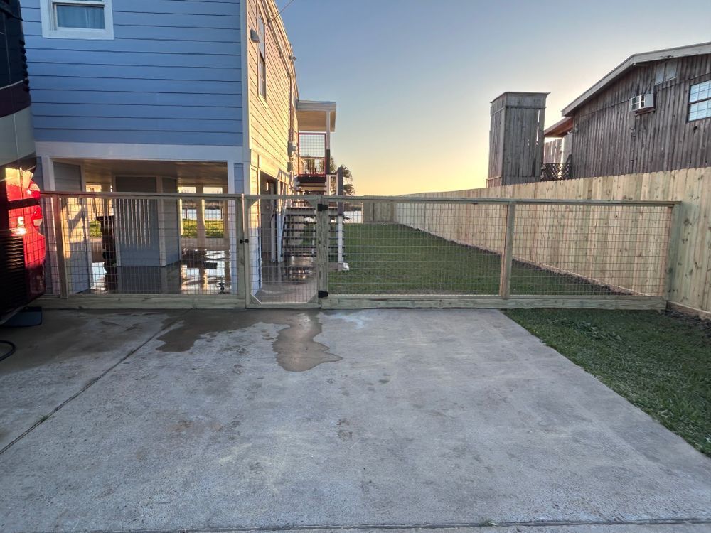 Fence with a gate surrounding a grassy yard, next to a light blue house and a concrete driveway.