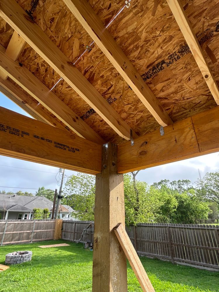 Wooden frame of a patio roof under construction with exposed beams, in a backyard with green grass and a fence.
