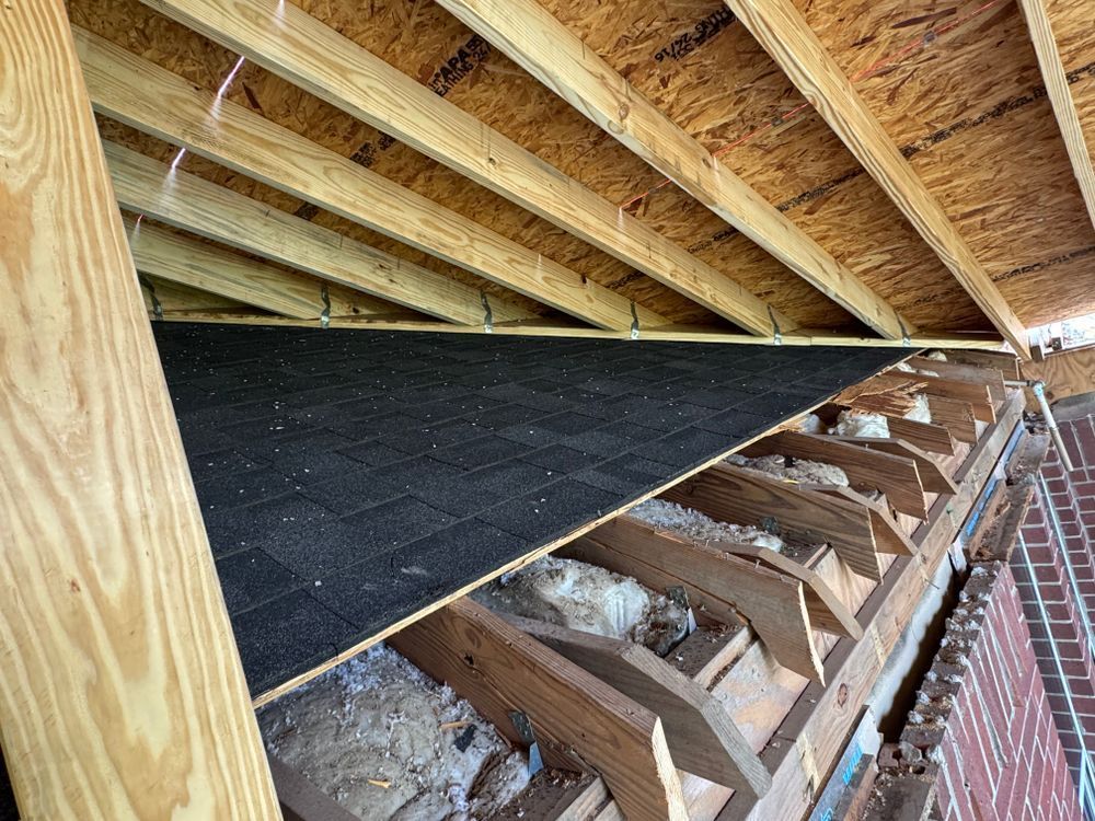 Interior roof view: wooden beams, black underlayment, brick wall, insulation, and plywood.