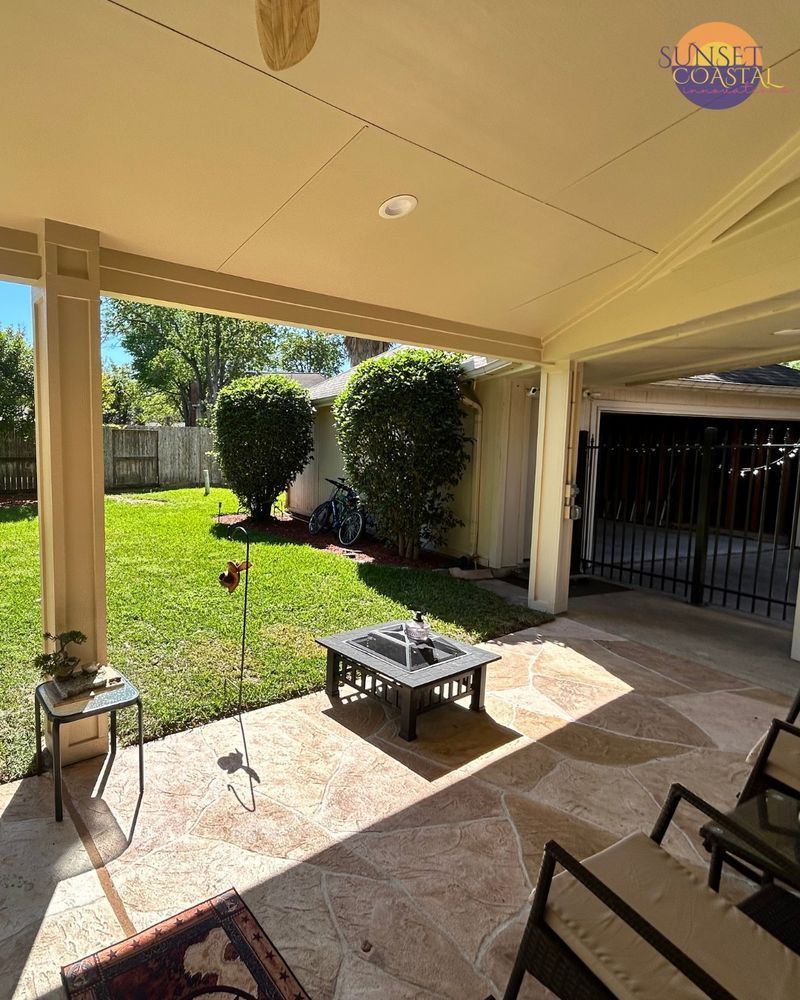 Covered patio with stone floor, overlooking a grassy backyard, fire pit, and small trees.