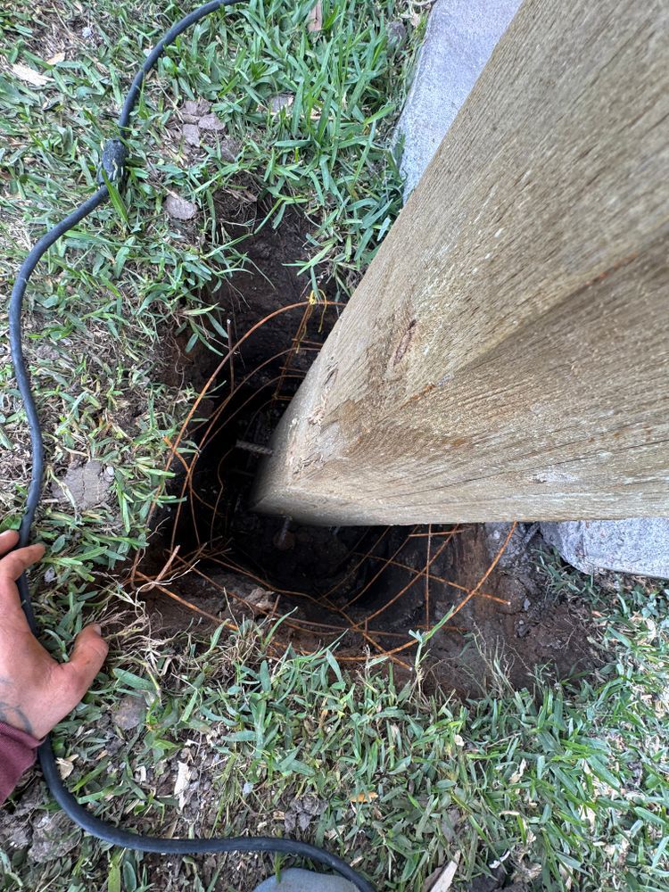 Hole in the ground with roots and a wooden post. A person's hand and an electrical cord are also visible.
